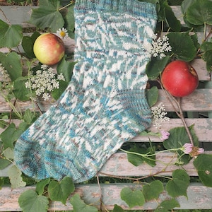 May include: A hand-knitted sock with a green, blue, and white patterned design. The sock is lying on a wooden bench with green leaves and red apples.