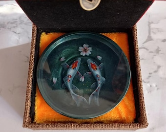 May include: A small, round, green dish with a clear top, containing a hand-painted image of two koi fish swimming in a pond with a white flower. The dish is in a small, square, brown box with a red velvet lining.