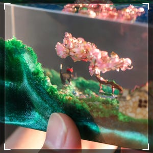 May include: A miniature diorama encased in clear resin, featuring a pink cherry blossom tree, a small house, and green foliage. The diorama is held in a hand.