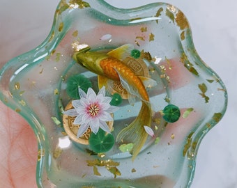 May include: A clear, star-shaped resin dish with a detailed miniature scene. It features a golden koi fish, a white and pink lotus flower, lily pads, and gold leaf accents. The dish is approximately 4 inches in diameter.