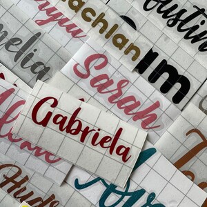 May include: A close-up of several sheets of adhesive vinyl with various names written on them in different colors and fonts. The names include "Lachlan", "Austin", "Sarah", "Gabriela", "Olivia", "Hudson", and "Ava".