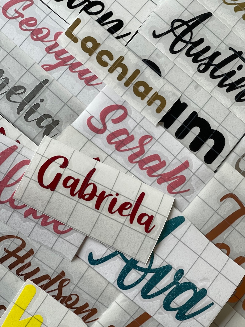 May include: A close-up of several sheets of adhesive vinyl with various names written on them in different colors and fonts. The names include "Lachlan", "Austin", "Sarah", "Gabriela", "Olivia", "Hudson", and "Ava".