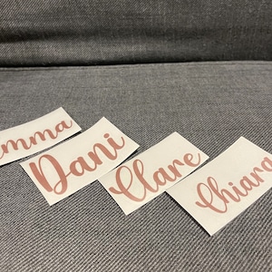 May include: Four white labels with the names Emma, Dani, Clare, and Chianna written in cursive script in a rose gold color.
