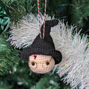 Puede incluir: Adorno navideño de ganchillo hecho a mano de un personaje con sombrero negro y gafas redondas. El adorno tiene un tono de piel claro y una cicatriz roja en forma de rayo en la frente. Cuelga de una cuerda roja, blanca y verde.