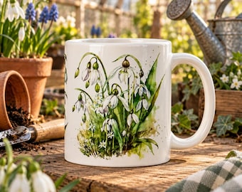 May include: A white ceramic mug featuring a detailed watercolor illustration of white snowdrop flowers with green stems and leaves. The design displays multiple blooming snowdrops and buds against a soft green wash background.