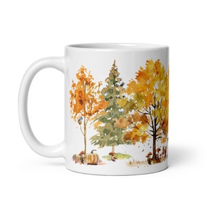Peut inclure: Mug en céramique blanche avec une illustration aquarelle d'arbres d'automne et une citrouille. Les arbres sont dans des tons de jaune, d'orange et de vert. La citrouille est orange.