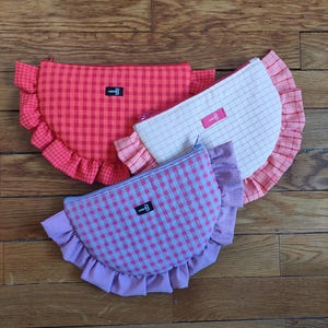 Op de afbeelding: Drie halfronde etuis met ritssluiting en gerimpelde randen. De etuis zijn in tinten rood, roze en lavendel, elk met een ruitpatroon. Elke etui heeft een klein label met een logo.