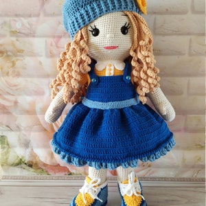 May include: A crocheted doll with blonde curly hair, wearing a blue dress, a blue beret with a yellow flower, and yellow and blue shoes.
