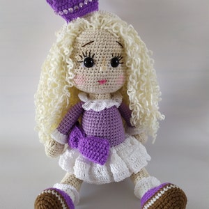 May include: A crocheted doll with blonde curly hair, wearing a purple dress, a white skirt, a purple bow, and a purple crown with rhinestones. The doll is sitting with its legs crossed and has brown shoes.