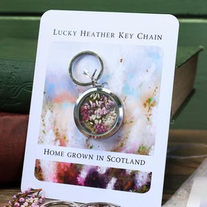Pode incluir: Um chaveiro Lucky Heather prateado com um pingente redondo de vidro preenchido com flores rosa e brancas secas. O cart&atilde;o diz "Home Grown in Scotland".