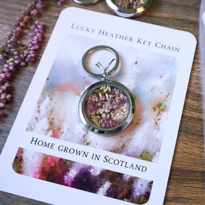 Pode incluir: Um chaveiro Lucky Heather prateado com um pingente redondo transparente preenchido com flores roxas e brancas secas. O chaveiro est&aacute; em um cart&atilde;o com o texto "Lucky Heather Key Chain" e "Home Grown in Scotland."