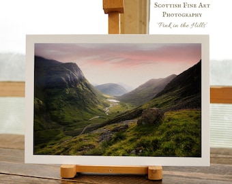Puede incluir: Una impresión enmarcada de un paisaje de valle con colinas verdes y un río, bajo un cielo rosa y naranja. La impresión se muestra en un pequeño caballete de madera. El texto en la impresión dice "Scottish Fine Art Photography 'Pink in the Hills'" y "A4 SIGNED PRINT BY ARTIST | KRISTY ASHTON."