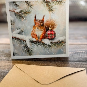 Pode incluir: Um esquilo vermelho est&aacute; sentado em um galho de pinheiro coberto de neve. O esquilo est&aacute; olhando para o espectador. Um enfeite de Natal vermelho e verde xadrez est&aacute; pendurado em um galho acima do esquilo.