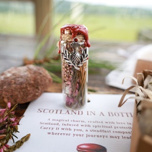 Puede incluir: Una peque&ntilde;a botella de vidrio llena de flores y hierbas secas, sellada con cera roja y un amuleto plateado con un cardo. La botella est&aacute; etiquetada como "Scotland in a Bottle" y la describe como un amuleto m&aacute;gico hecho con amor en Escocia, infundido con protecci&oacute;n espiritual.