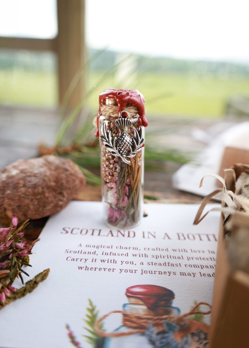 Puede incluir: Una peque&ntilde;a botella de vidrio llena de flores y hierbas secas, sellada con cera roja y un amuleto plateado con un cardo. La botella est&aacute; etiquetada como "Scotland in a Bottle" y la describe como un amuleto m&aacute;gico hecho con amor en Escocia, infundido con protecci&oacute;n espiritual.
