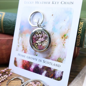 Pode incluir: Um porta-chaves prateado com um berloque redondo e transparente preenchido com flores roxas e brancas secas. O porta-chaves &eacute; exibido em um cart&atilde;o que diz "Lucky Heather Key Chain" e "Home Grown in Scotland".