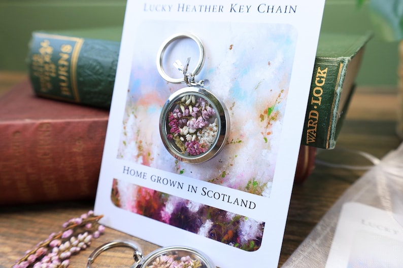 Pode incluir: Um porta-chaves prateado com um berloque redondo e transparente preenchido com flores roxas e brancas secas. O porta-chaves &eacute; exibido em um cart&atilde;o que diz "Lucky Heather Key Chain" e "Home Grown in Scotland".