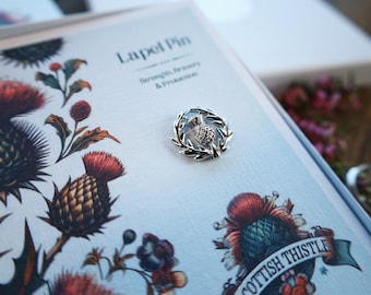May include: A silver-toned lapel pin shaped like a thistle, resting on a light-colored card featuring botanical illustrations of thistles and the text Lapel Pin, Strength, Bravery & Protection, and Scottish Thistle.