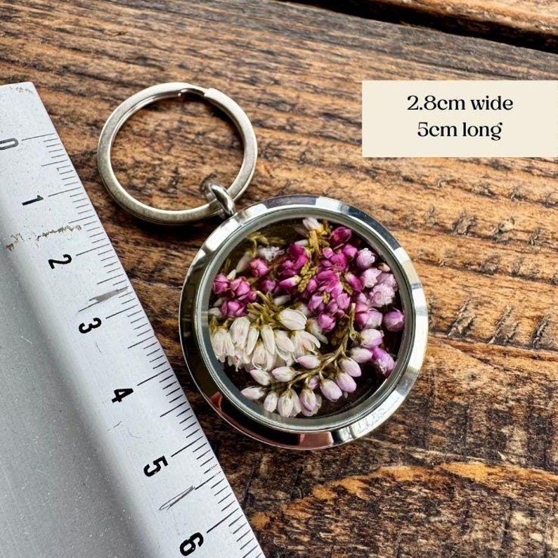 Pode incluir: Um pingente de chaveiro redondo prateado preenchido com flores secas roxas e brancas. O pingente tem 2,8 cm de largura e 5 cm de comprimento. O fundo &eacute; uma superf&iacute;cie de madeira. Um acess&oacute;rio decorativo.