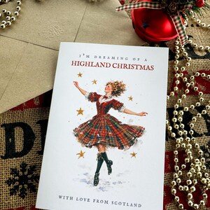 Pu&ograve; includere: Un biglietto d'auguri bianco con il testo I'M DREAMING OF A HIGHLAND CHRISTMAS e WITH LOVE FROM SCOTLAND. L'illustrazione mostra una persona con un abito a quadri rosso e verde che danza tra stelle dorate, con una busta abbinata accanto.