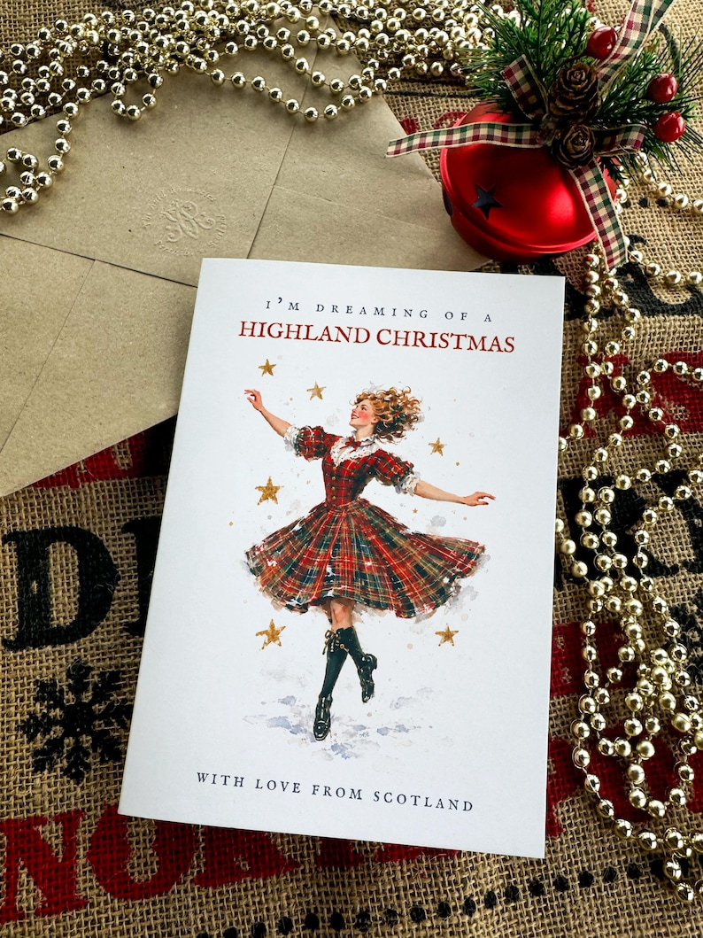 Pu&ograve; includere: Un biglietto d'auguri bianco con il testo I'M DREAMING OF A HIGHLAND CHRISTMAS e WITH LOVE FROM SCOTLAND. L'illustrazione mostra una persona con un abito a quadri rosso e verde che danza tra stelle dorate, con una busta abbinata accanto.