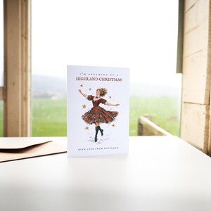 Pu&ograve; includere: Un biglietto d'auguri bianco con un'illustrazione centrale di una persona che danza in un abito scozzese rosso e verde con stivali neri, circondata da stelle dorate. Il testo recita I'M DREAMING OF A HIGHLAND CHRISTMAS e WITH LOVE FROM SCOTLAND.