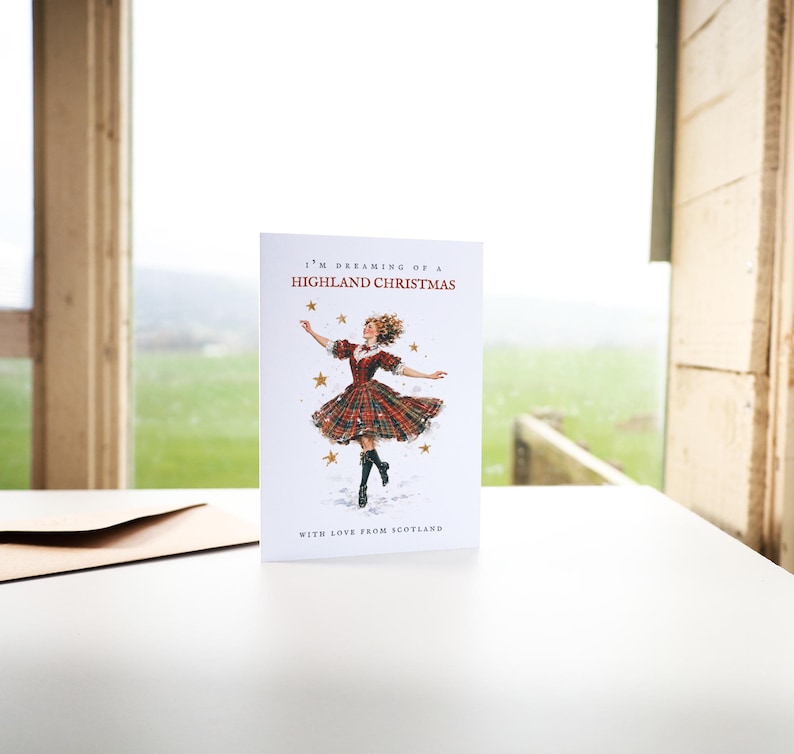 Pu&ograve; includere: Un biglietto d'auguri bianco con un'illustrazione centrale di una persona che danza in un abito scozzese rosso e verde con stivali neri, circondata da stelle dorate. Il testo recita I'M DREAMING OF A HIGHLAND CHRISTMAS e WITH LOVE FROM SCOTLAND.