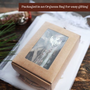 Puede incluir: Una peque&ntilde;a caja marr&oacute;n con una ventana transparente, mostrando un arreglo de flores secas. La caja est&aacute; empaquetada en una bolsa de organza blanca, lo que la hace ideal para regalar.