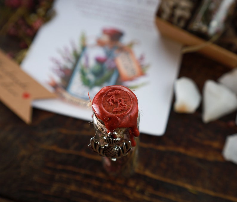 Puede incluir: Una peque&ntilde;a botella de vidrio con un sello de cera roja y un amuleto de plata. El sello de cera tiene un dise&ntilde;o de una letra 'R' estilizada.