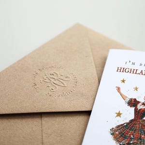 Pu&ograve; includere: Una busta in carta kraft marrone con un logo circolare in rilievo e un biglietto d'auguri bianco con un'illustrazione ad acquerello di una persona in abito a quadri rossi con stelle dorate e il testo I'M DREAMING OF A HIGHLAND CHRISTMAS.