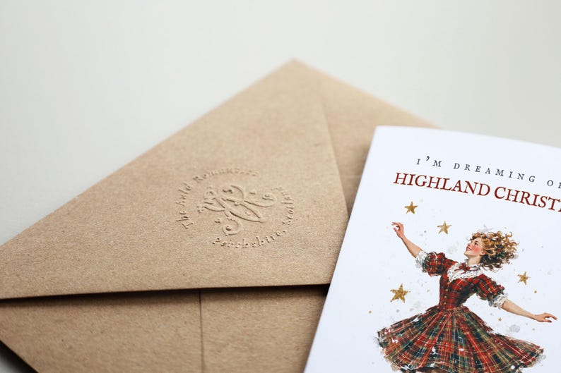 Pu&ograve; includere: Una busta in carta kraft marrone con un logo circolare in rilievo e un biglietto d'auguri bianco con un'illustrazione ad acquerello di una persona in abito a quadri rossi con stelle dorate e il testo I'M DREAMING OF A HIGHLAND CHRISTMAS.