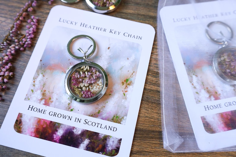 Pode incluir: Um chaveiro Lucky Heather prateado com um pingente redondo transparente preenchido com flores roxas e brancas secas. O chaveiro est&aacute; em um cart&atilde;o com o texto "Lucky Heather Key Chain" e "Home Grown in Scotland."
