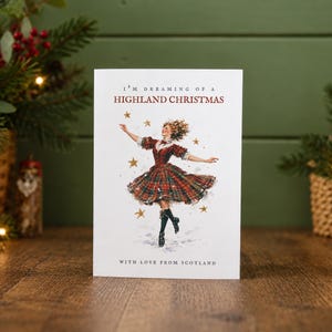 Pu&ograve; includere: Un biglietto d'auguri bianco con l'illustrazione di una persona che danza indossando un abito scozzese rosso e verde e stivali neri. Il testo recita I'M DREAMING OF A HIGHLAND CHRISTMAS e WITH LOVE FROM SCOTLAND, circondato da stelle dorate.