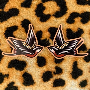 May include: Two stickers featuring a traditional tattoo-style swallow design. The birds are black with red and gold accents, outlined in black, and set against a peach-colored background. The background is a leopard print.