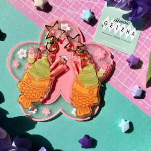 Op de afbeelding: Een paar hangende oorbellen met groen matcha-ijs in een taiyaki-visvormige wafeltje met een roze kers op de top. De oorbellen zijn omringd door roze kersenbloesems en gouden sterren.
