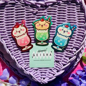 May include: Three die-cut stickers featuring cartoon characters inside colorful drinks with cherries and straws. The left sticker is pink, the center is green, and the right is blue. A square card with the text Atomic Geisha Design is included.