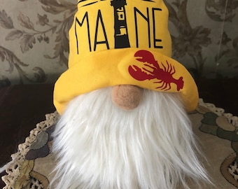 May include: A yellow gnome hat with the word "MAINE" and a lobster design. The gnome has a white beard and a brown nose.