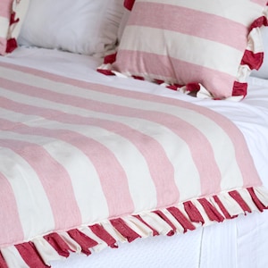 Puede incluir: Una cama con una colcha blanca y dos almohadas. La colcha y las almohadas presentan un patrón de rayas rosas y blancas. Las almohadas tienen un ribete de volantes rojos, y la colcha tiene un ribete de volantes rojos en el borde inferior.