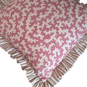 May include: Square decorative pillow with a white background and a repeating pattern of pink coral-like shapes. The pillow has a ruffled edge with a striped pattern in shades of cream and brown. A cozy home decor item.