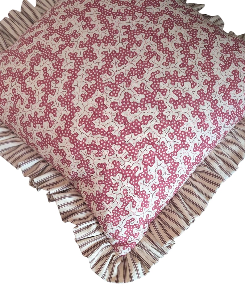 May include: Square decorative pillow with a white background and a repeating pattern of pink coral-like shapes. The pillow has a ruffled edge with a striped pattern in shades of cream and brown. A cozy home decor item.