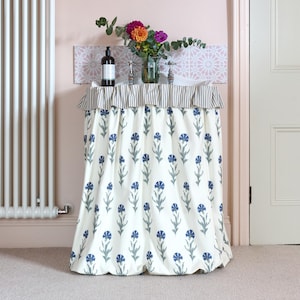 May include: A white sink skirt with a blue floral pattern. The skirt is draped over a sink with silver faucets and a striped ruffle. A bottle of soap and a vase of flowers sit on the sink's surface.