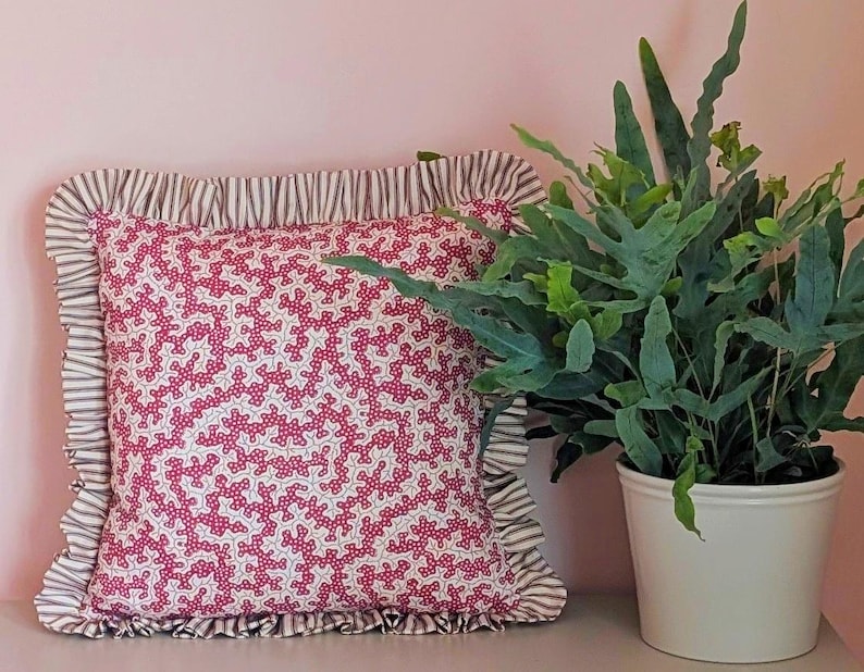 May include: A square decorative pillow with a white background and a red floral pattern. The pillow has a ruffled edge with a striped pattern. A potted fern with green leaves sits to the right of the pillow.