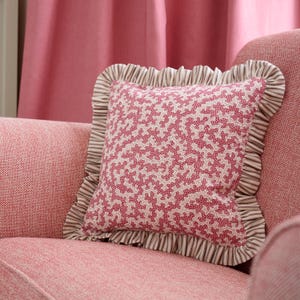 May include: A square decorative pillow with a coral-like pattern in shades of red and cream. The pillow has a ruffled border with a striped pattern in cream and brown. The pillow is placed on a pink upholstered chair.