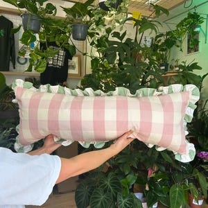 Könnte beinhalten: Ein rechteckiges Kissen mit rosa-weißem Vichy-Muster. Es hat einen gerüschten Rand in Hellgrün. Das Kissen wird vor einem Hintergrund aus Pflanzen und hängenden Töpfen präsentiert, was eine natürliche Umgebung schafft.