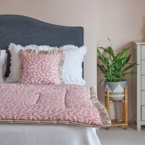 May include: A bed with a gray headboard, white bedding, and a red and white patterned quilt and pillows. A gray dresser and a plant in a woven pot are in the background.