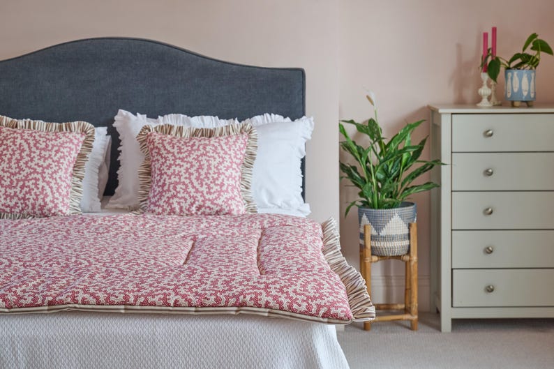 May include: A bed with a gray headboard, white bedding, and a red and white patterned quilt and pillows. A gray dresser and a plant in a woven pot are in the background.