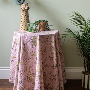 May include: A round table covered with a pink floral tablecloth. A tall, tan leopard-shaped vase, a green glass, and a small potted plant sit on the table. A large potted plant is to the right.