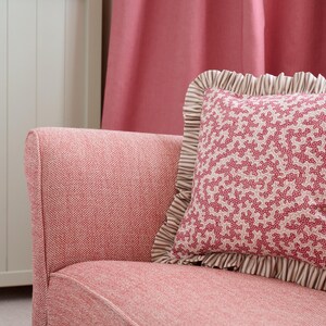 May include: A pink armchair with a patterned pillow. The pillow has a coral-like design in red and white, framed by a ruffled edge with brown and white stripes. The chair and pillow create a cozy, inviting aesthetic.