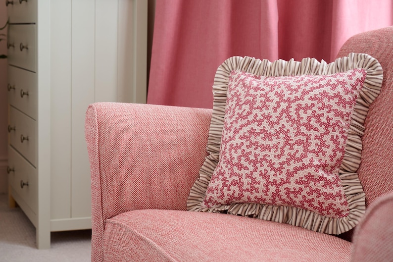 May include: A pink armchair with a patterned pillow. The pillow has a coral-like design in red and white, framed by a ruffled edge with brown and white stripes. The chair and pillow create a cozy, inviting aesthetic.
