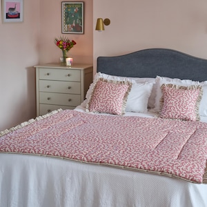 May include: A bed with a gray headboard, white bedding, and a red and white patterned quilt. Two matching throw pillows with ruffled edges sit on top. The room has pink walls and decorative wall art.